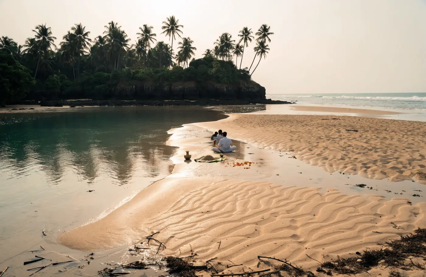 A wide pale sandbar at low tide near Gokarna where a slow river meets the Arabian Sea, a small family seated at the water's edge with a priest, a brass vessel, a banana leaf of rice and loose marigold petals on the wet sand