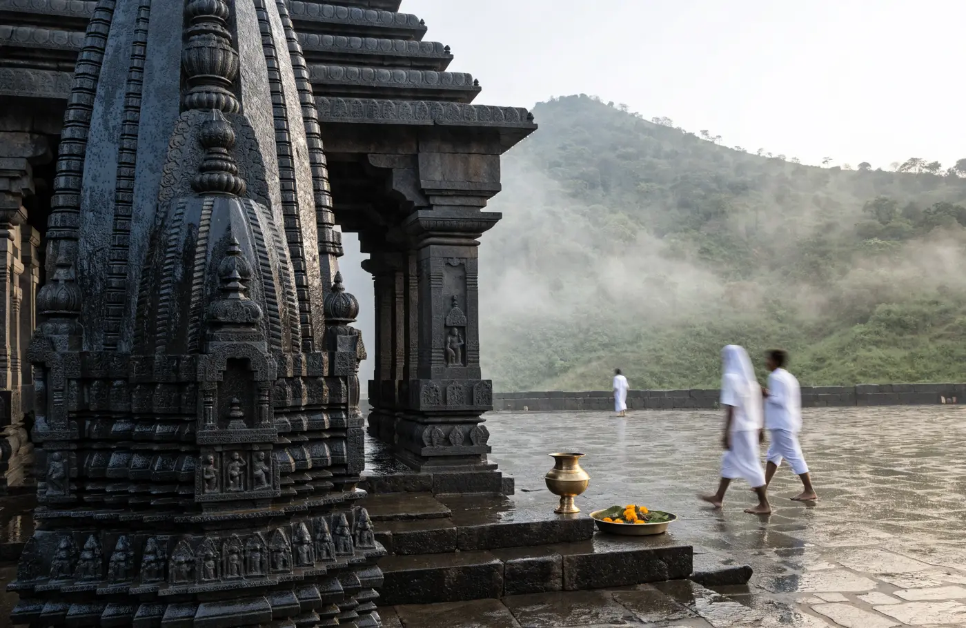 The carved black basalt shikhara of the Trimbakeshwar temple after rain, a brass kalash and a thali of marigolds on the step, the green slopes of Brahmagiri hill rising in mist behind