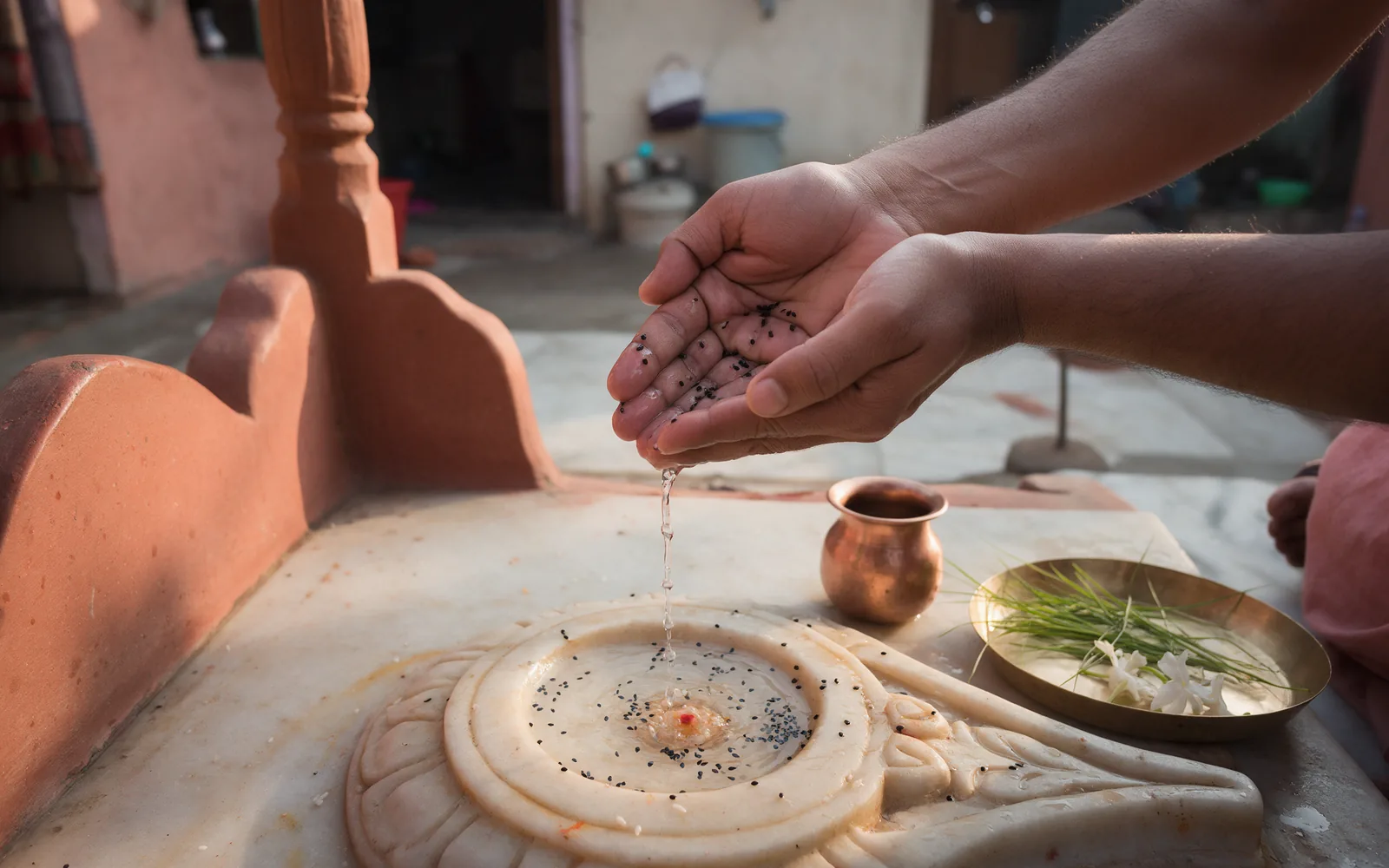Cupped bare hands releasing a thin stream of water mixed with black sesame seeds onto a carved marble tulsi platform in a home courtyard, a copper lota and a brass thali of kusha grass and white flowers beside them in early morning light