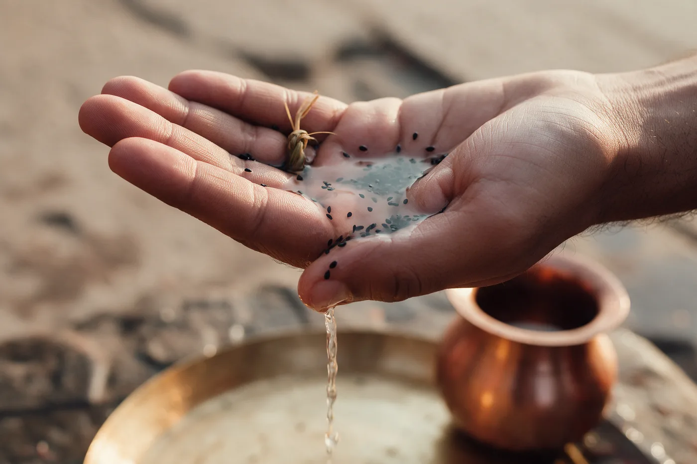 Macro close-up of an open palm cupping water and black sesame seeds, the water draining off the thumb edge of the hand in a thin stream into a brass plate below, a ring of kusha grass on the finger