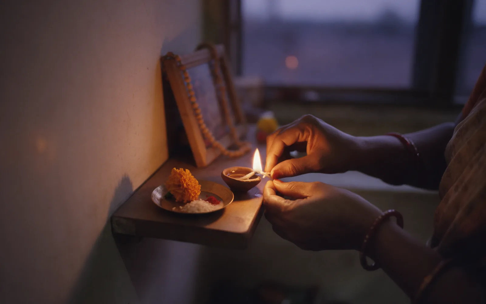 Hands lighting a small clay oil lamp at dusk on a wooden home altar shelf, beside marigolds and a garlanded photo frame, evening light at the window behind